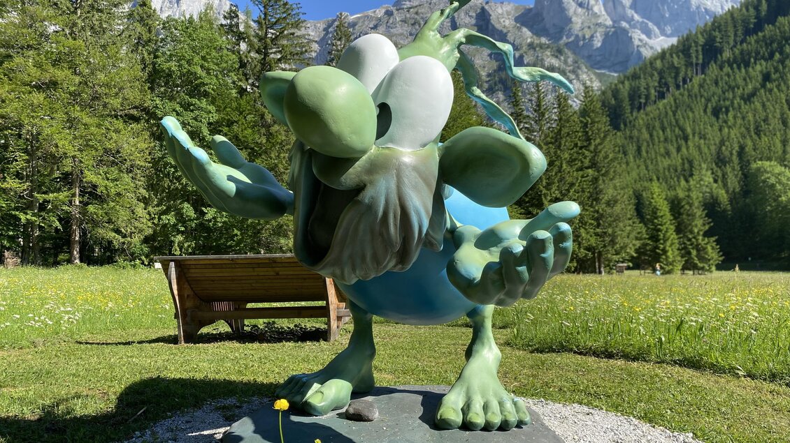 Eine große, bunte Statue eines phantastischen Wesens steht in einer Wiese. Im Hintergrund sind Berge und Bäume zu sehen.