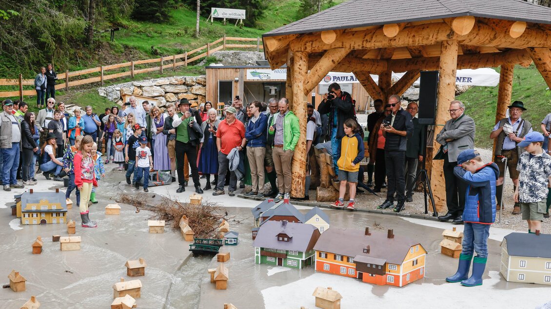Eine Gruppe von Menschen versammelt sich unter einem Holzpavillon, während sie einem Modell eines Dorfes mit kleinen Häusern und einem fließenden Wasserlauf zuschaut. Die Szene spielt sich in einer grünen, waldreichen Umgebung ab. | © Martin Huber
