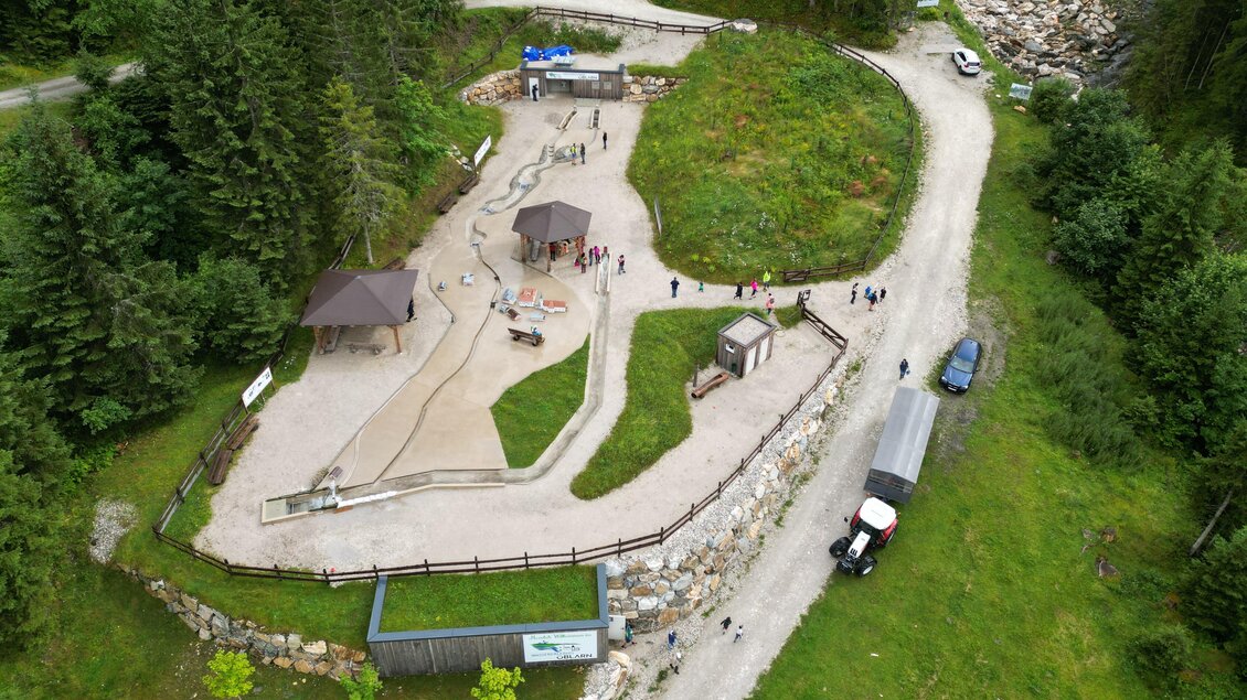 Ein Spielplatz in einer grünen Umgebung mit einem Spielbereich aus Sand und Wasser. Im Hintergrund sind Bäume und eine Straße zu sehen. | © Wassererlebnis Öblarn