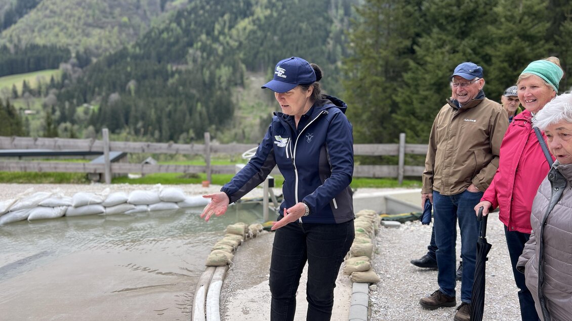 Eine Person erklärt etwas am Ufer eines Gewässers. Im Hintergrund sind grüne Hügel und Bäume zu sehen, während einige Zuschauer aufmerksam zuschauen. | © Wassererlebnis Öblarn