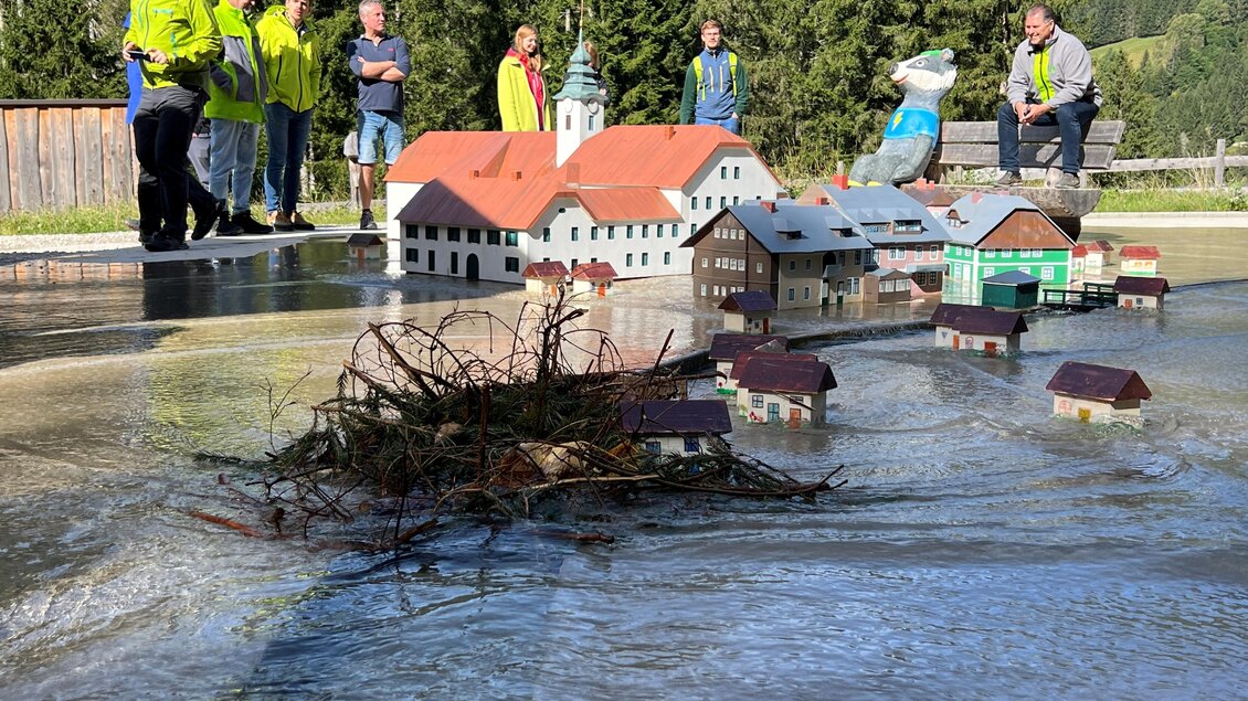 Eine Gruppe von Personen steht neben einer Miniaturstadt, die im Wasser schwimmt. Im Vordergrund sind Äste und kleine Häusermodelle sichtbar. | © Natalie Prüggler