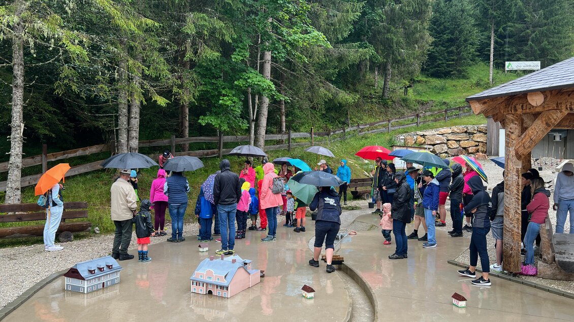 Eine Gruppe von Menschen mit Regenschirmen steht in einem nassen Bereich im Freien. Im Hintergrund sind Bäume und Miniaturhäuser zu sehen. | © Wassererlebnis Öblarn