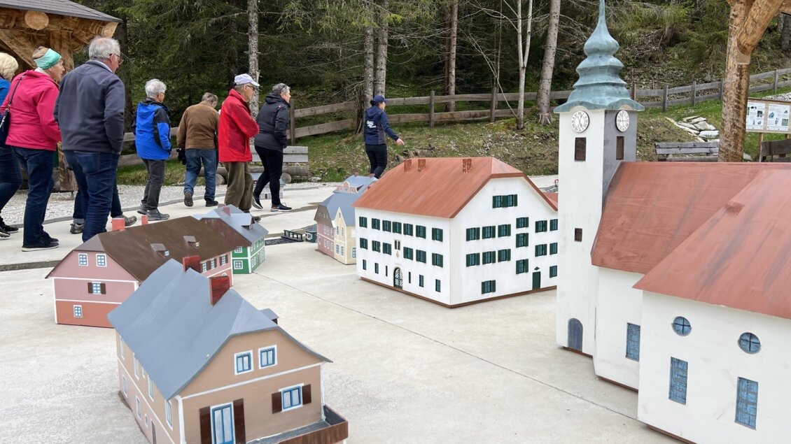 Eine Gruppe von Menschen spaziert vorbei an Miniaturmodellen von Häusern und einer Kirche. Im Hintergrund sind Bäume und eine natürliche Umgebung zu sehen. | © Wassererlebnis Öblarn