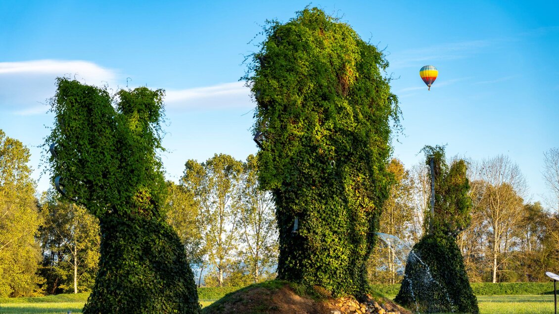 Eine grüne Skulptur aus Pflanzenform, die drei Hasen darstellt, steht in einer Wiese. Im Hintergrund fliegt ein bunter Heißluftballon am blauen Himmel. | © Kurkommission Bad Blumau