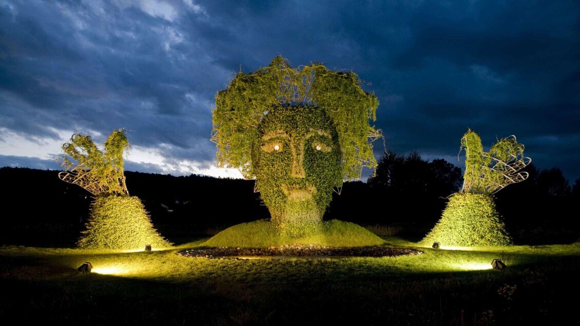 Eine große, grüne Skulptur eines Gesichts mit Händen aus Pflanzen vor einem bewölkten Himmel. Die Figur wird nachts von Lichtern beleuchtet. | © Rogner Bad Blumau