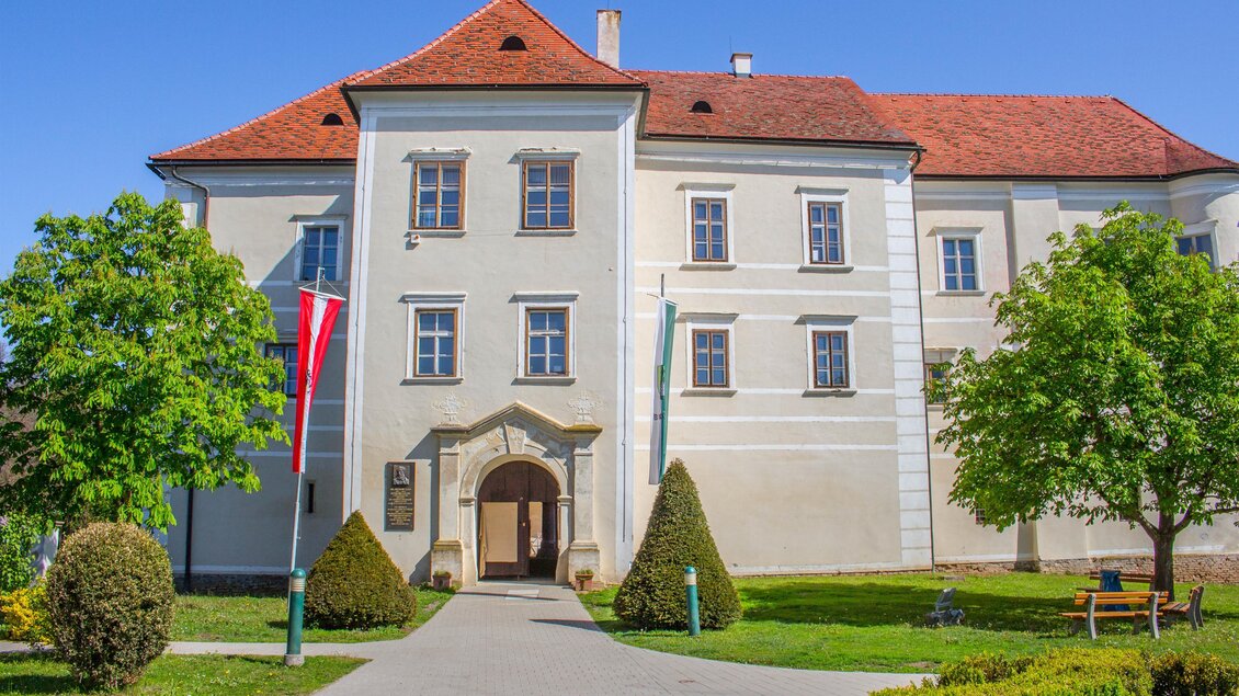 Ein historisches Gebäude mit roten Dächern und großen Fenstern. Der Eingang ist von gepflegten Bäumen und Sträuchern umgeben. | © Sandra Brünner