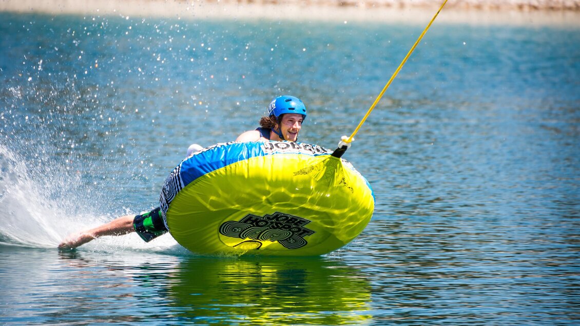 Ein Sportler fährt auf einem gelben Wasserschlauch über einen klaren See. Spritzwasser fliegt hoch, während er mit viel Schwung und Freude fährt. | © www.mariazell.blog | Fred Lindmoser
