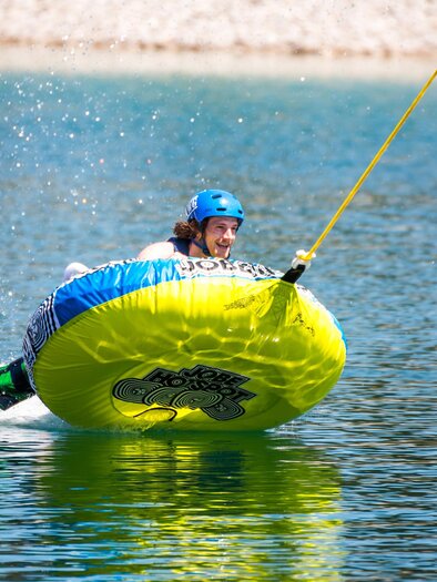 An athlete rides on a yellow water tube over a clear lake. Spray water flies high as he rides with a lot of momentum and joy. | © www.mariazell.blog | Fred Lindmoser