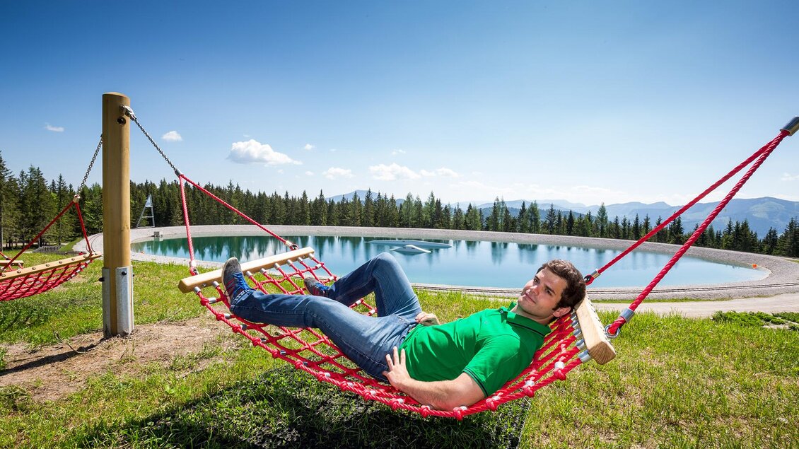 Ein Mann liegt entspannt in einer roten Hängematte auf einer Wiese. Im Hintergrund ist ein klarer See und eine bewaldete Landschaft zu sehen. | © www.mariazell.blog | Fred Lindmoser