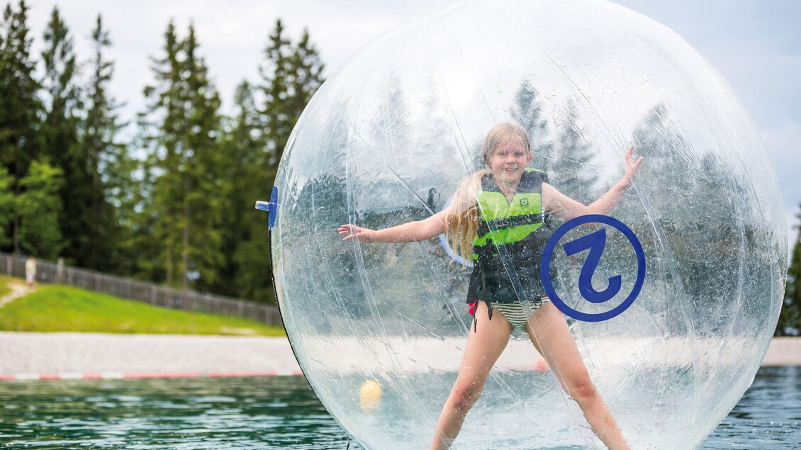 Ein Mädchen steht in einer transparenten Wasserblase auf einem See. Sie trägt eine Schwimmweste und hat Spaß im Wasser.