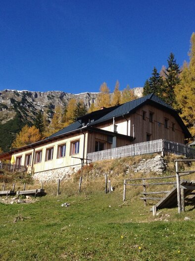 A house in the mountains surrounded by green meadows and autumn trees. In the background, majestic mountains are visible.