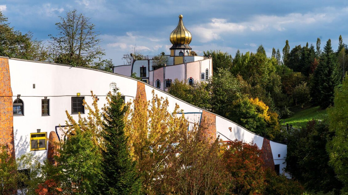 Eine ungewöhnliche Gebäudeansicht mit einer goldenen Kuppel und bunten Bäumen im Vordergrund. Der Himmel ist bewölkt, was eine ruhige Atmosphäre schafft. | © Kurkommission Bad Blumau