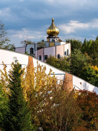Eine ungewöhnliche Gebäudeansicht mit einer goldenen Kuppel und bunten Bäumen im Vordergrund. Der Himmel ist bewölkt, was eine ruhige Atmosphäre schafft. | © Kurkommission Bad Blumau