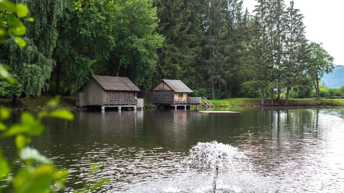 Eine ruhige Landschaft mit einem Teich und zwei Holzchalets. Im Wasser sprudelt ein Wasserfall, umgeben von grünen Bäumen. | © Anita Fössl