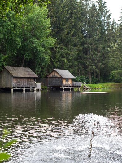 A peaceful landscape with a pond and two wooden chalets. In the water, a waterfall bubbles, surrounded by green trees. | © Anita Fössl