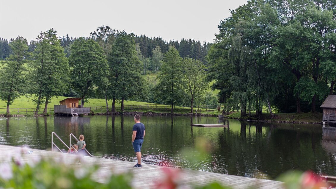 Ein ruhiger See umgeben von Bäumen und Wiesen. Eine Person spaziert entlang der Uferpromenade. | © Anita Fössl