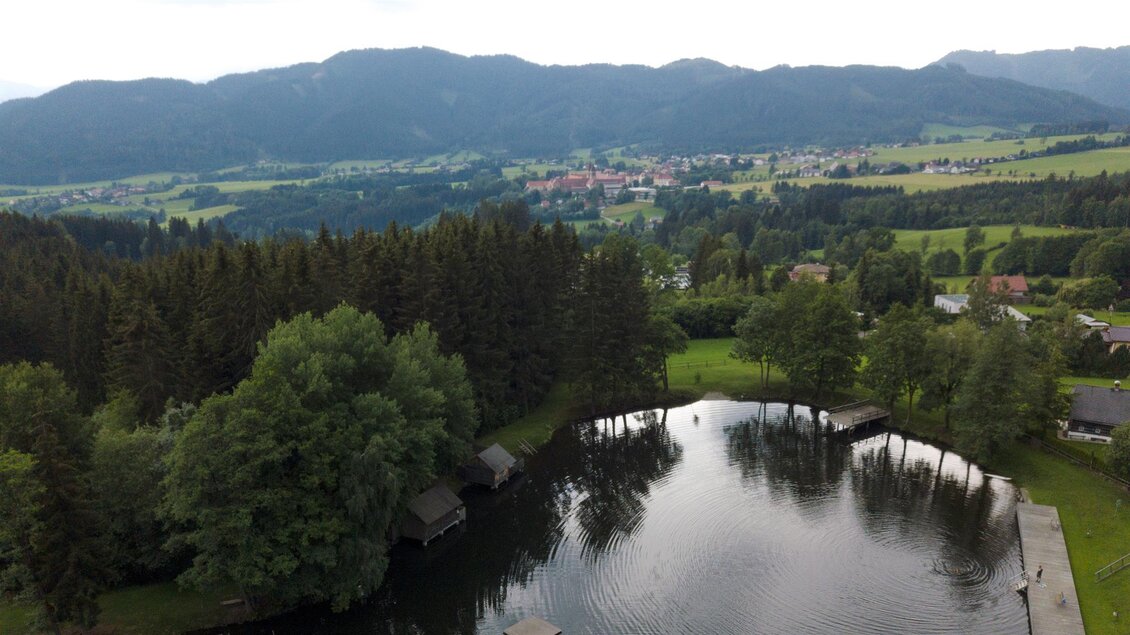 Eine malerische Landschaft mit einem ruhigen See und dichtem Wald. Im Hintergrund sind sanfte Hügel und ein Dorf zu sehen. | © Anita Fössl