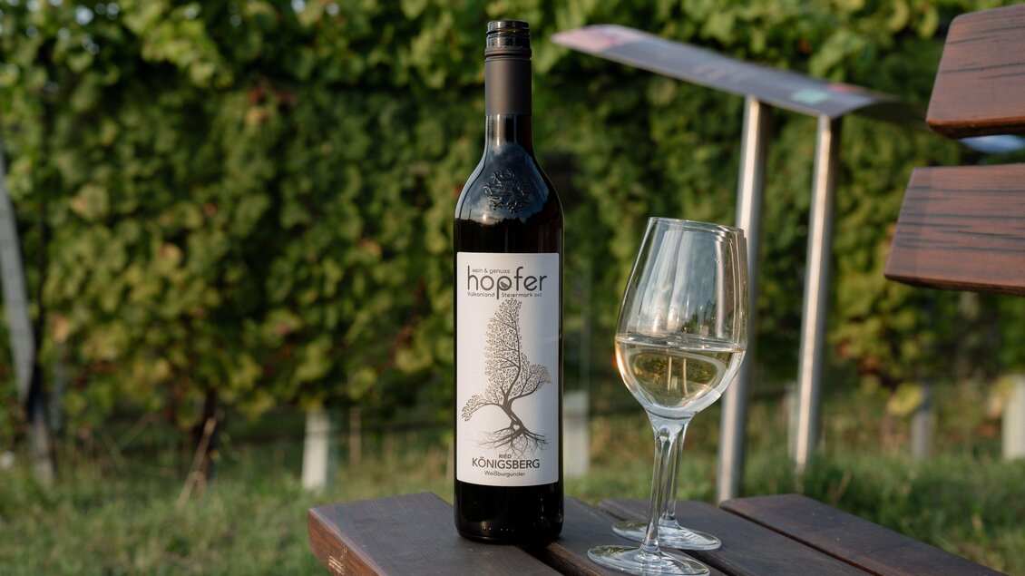 Eine Flasche Wein steht neben einem Weinglas auf einem Tisch. Im Hintergrund sind Weinreben zu sehen.