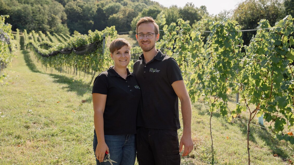 Ein junges Paar steht in einem Weinberg zwischen Reihen von Weinreben. Die Landschaft ist grün und die Sonne scheint. | © Weingut Hopfer