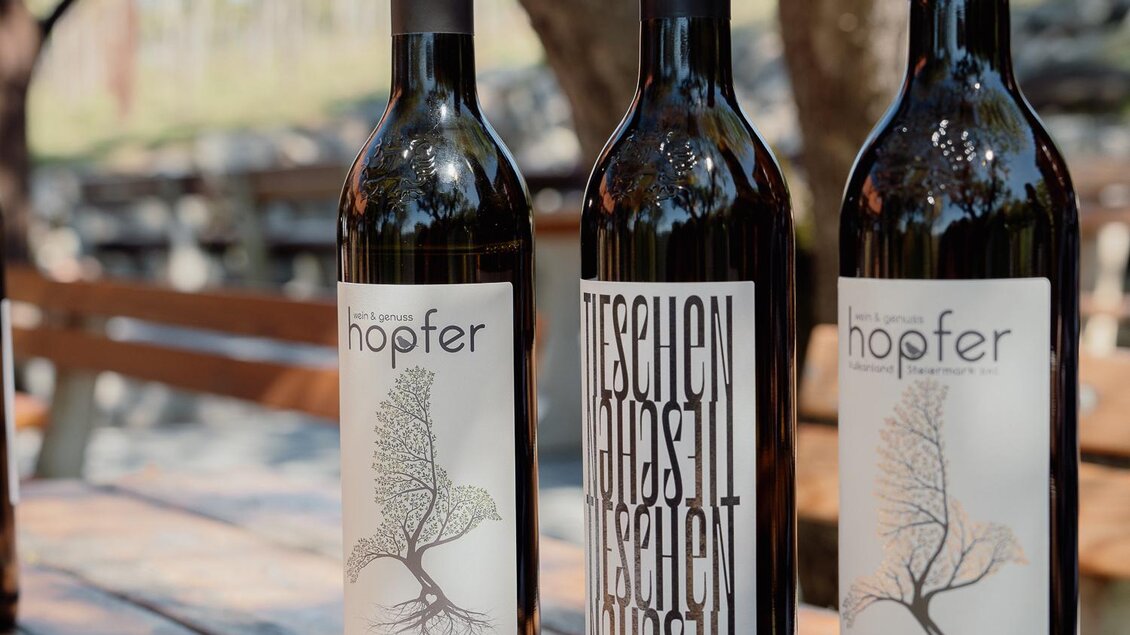 Drei Flaschen Wein mit ansprechenden Etiketten stehen auf einem Holztisch. Im Hintergrund sieht man Bäume und eine weite, grüne Landschaft. | © Weingut Hofper