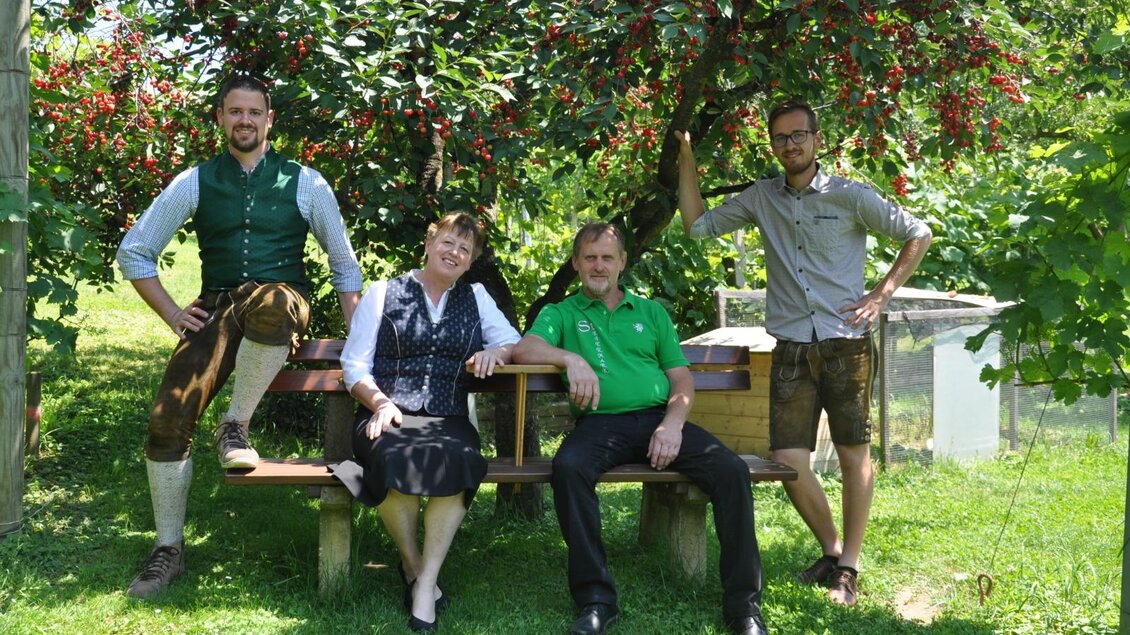Eine Gruppe von vier Personen sitzt unter einem Kirschbaum. Sie tragen traditionelle Kleidung und lächeln in die Kamera. | © Weingut Hopfer