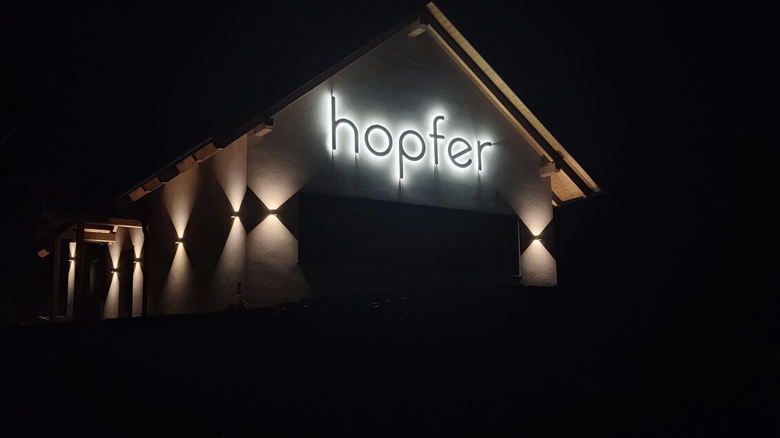 Ein modernes Gebäude mit beleuchtetem Schild, das "hopfer" zeigt. Die Umgebung ist dunkel, wodurch das Licht des Schildes auffällt.