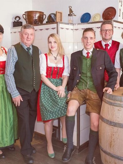 Eine Gruppe von Menschen in traditioneller bayerischer Tracht steht in einem gemütlichen Raum. Im Hintergrund ist ein Kamin zu sehen, und sie lächeln freundlich in die Kamera. | © Netwerker