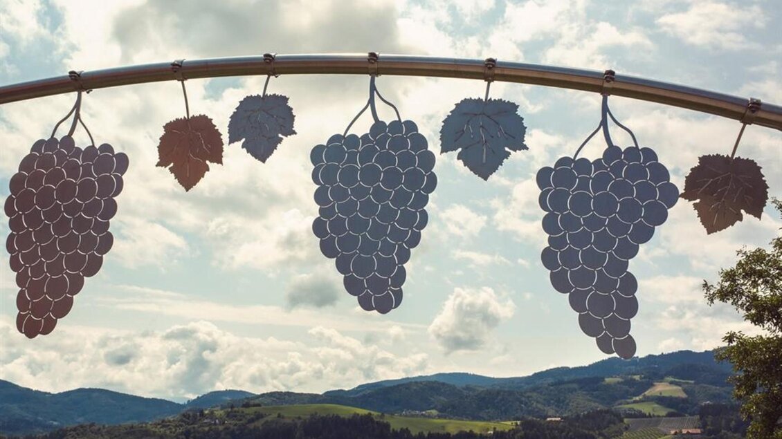 Ein kreatives Kunstwerk mit stilisierten Trauben hängt über einer grünen Landschaft. Im Hintergrund sind sanfte Hügel und ein bewölkter Himmel zu sehen. | © Netwerker
