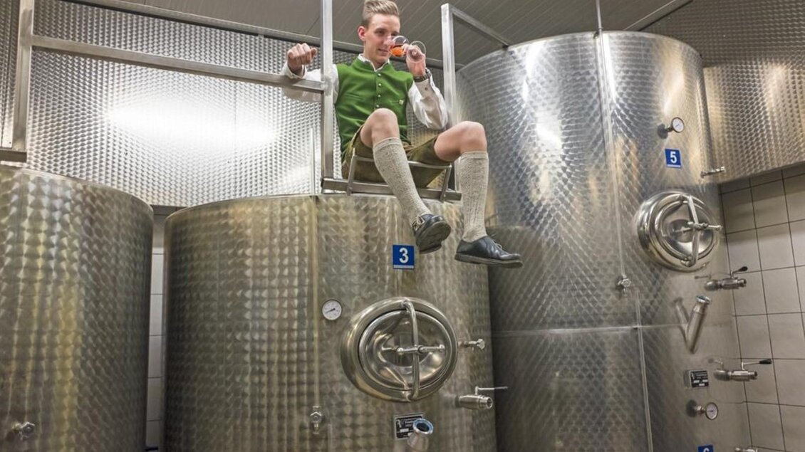 Ein Mann sitzt auf einem großen Metalltank in einer Brauerei. Er trägt traditionelle Kleidung und genießt ein Getränk. | © Netwerker