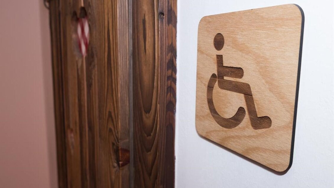 Ein Holzschild mit dem Symbol für barrierefreies WC. Es ist neben einer Holz- bzw. Wandtür angebracht. | © Netwerker
