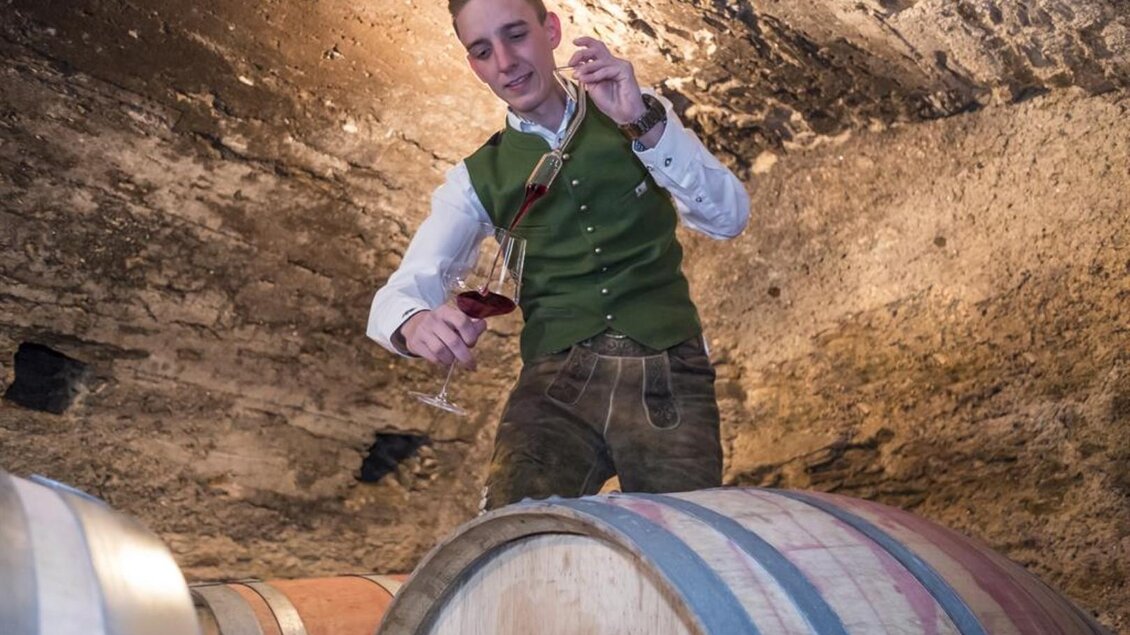 Ein Mann in traditioneller Kleidung steht auf einem Weinfass und schenkt Wein in ein Glas ein. Im Hintergrund sind weitere Holzfässer und die Wände einer Weinkellerei zu sehen. | © Netwerker