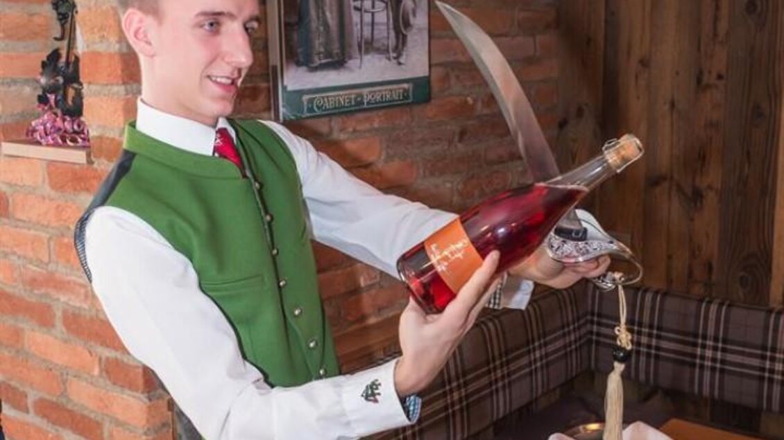 Ein Kellner in traditioneller Tracht öffnet eine Flasche Wein mit einem Säbel. Der Hintergrund zeigt eine gemütliche, rustikale Einrichtung. | © Netwerker