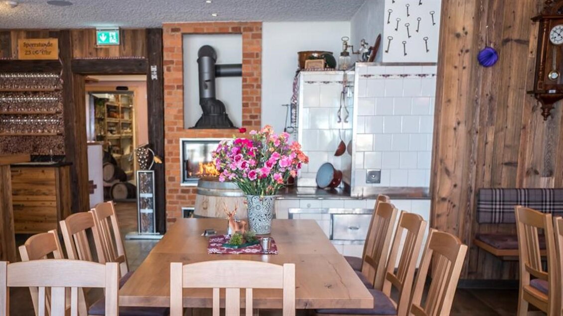 Ein gemütlicher Essbereich mit einem großen Holztisch und Stühlen. Auf dem Tisch steht eine Blumenvase und im Hintergrund sind Holzregale und eine traditionelle Küche zu sehen. | © Netwerker