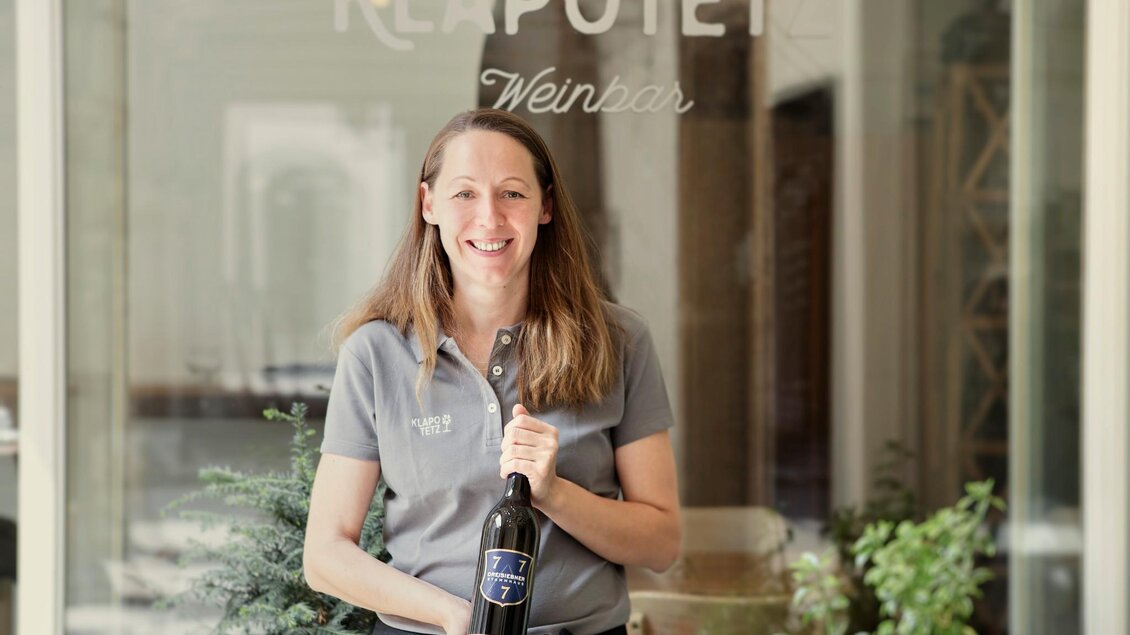 Eine lächelnde Frau hält eine Flasche Wein vor dem Schaufenster eines Weinladens. Im Hintergrund sind Pflanzen und der Schriftzug "Klapotetz" zu sehen. | © Klapotetz