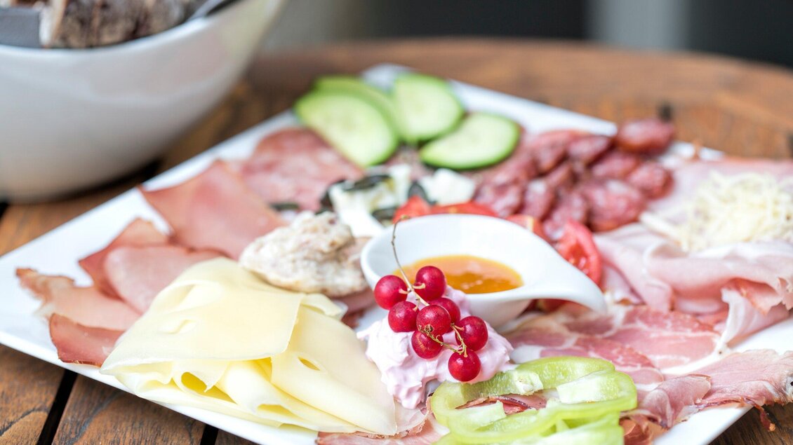 Ein Teller mit verschiedenen Wurst- und Käseaufschnitten, garniert mit Gurkenscheiben und Tomaten. In der Mitte befindet sich eine kleine Schale mit Dipsauce. | © Klapotetz