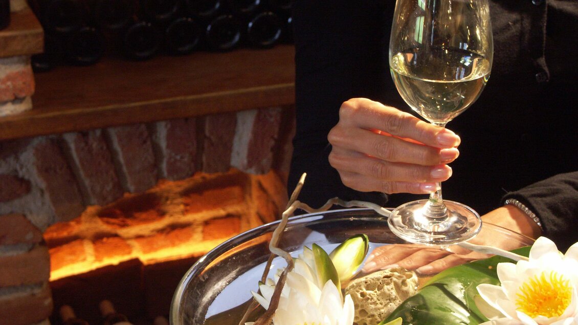 Ein elegantes Ambiente mit einem Glas Weißwein in einer Hand und hübschen Seerosen auf einem Tisch. Im Hintergrund ist eine Steinmauer mit Weinflaschen sichtbar. | © Rogner Bad Blumau