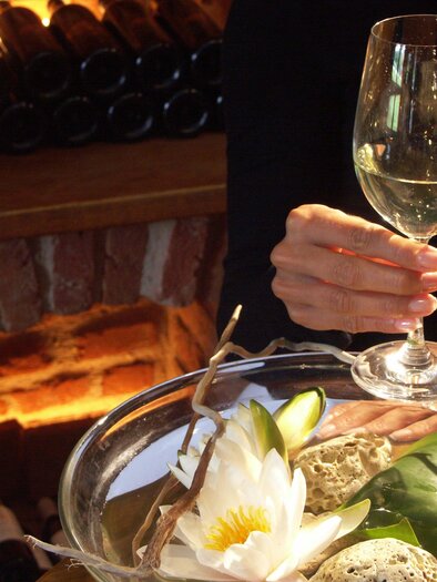 Ein elegantes Ambiente mit einem Glas Weißwein in einer Hand und hübschen Seerosen auf einem Tisch. Im Hintergrund ist eine Steinmauer mit Weinflaschen sichtbar. | © Rogner Bad Blumau
