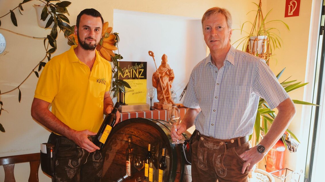 Zwei Männer stehen vor einem Weinfass und präsentieren Weinflaschen. Im Hintergrund ist eine Statue und eine Pflanze zu sehen. | © Lederer