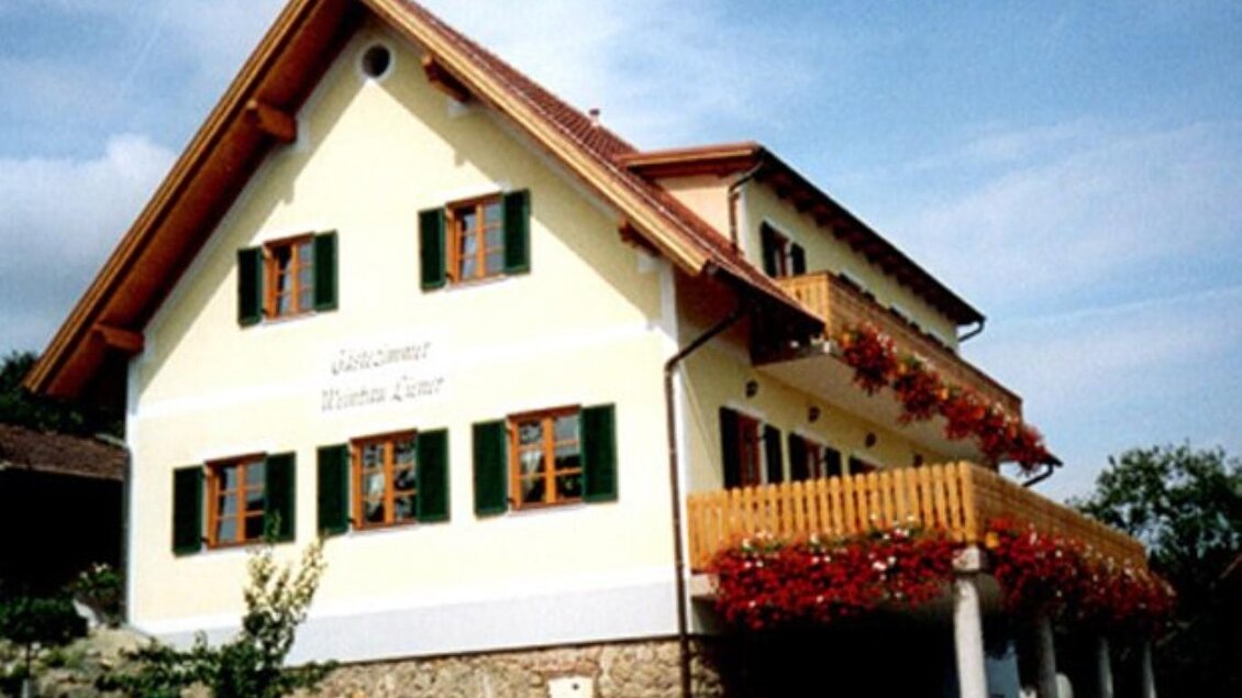 Ein zweistöckiges Haus mit einem hellen Anstrich und grünen Fensterläden. Der Balkon ist mit Blumen geschmückt und das Gebäude steht in einer grünen Umgebung. | © Liener