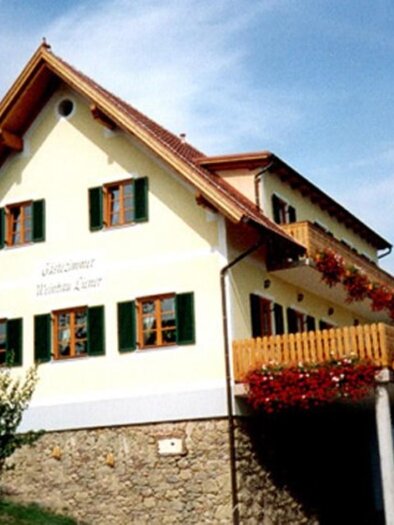 Ein zweistöckiges Haus mit einem hellen Anstrich und grünen Fensterläden. Der Balkon ist mit Blumen geschmückt und das Gebäude steht in einer grünen Umgebung. | © Liener