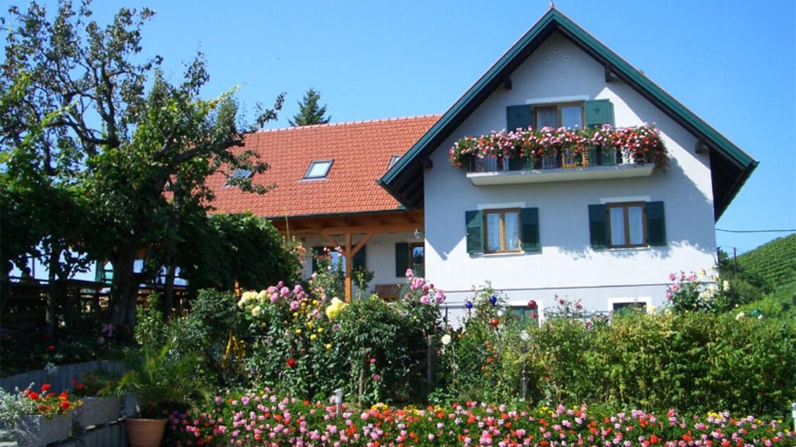 Ein hübsches Haus mit einem roten Dach und blauen Fensterläden. Der Garten ist üppig mit bunten Blumen und grünen Pflanzen gestaltet. | © Trunk