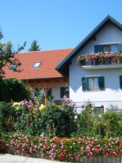 A beautiful house with a red roof and blue shutters. The garden is lush, designed with colorful flowers and green plants. | © Trunk
