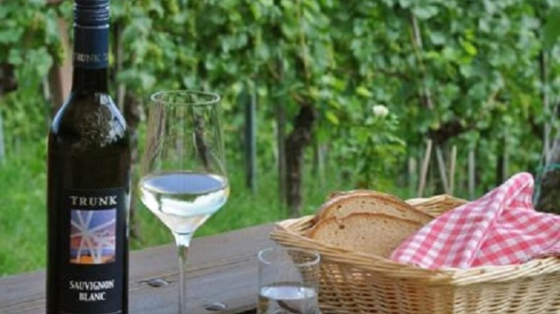 Ein Tisch im Freien mit einer Flasche Weißwein und einem Glas. Darauf liegt eine Platte mit verschiedenen Speisen und Brot, umgeben von Weinbergen. | © Trunk