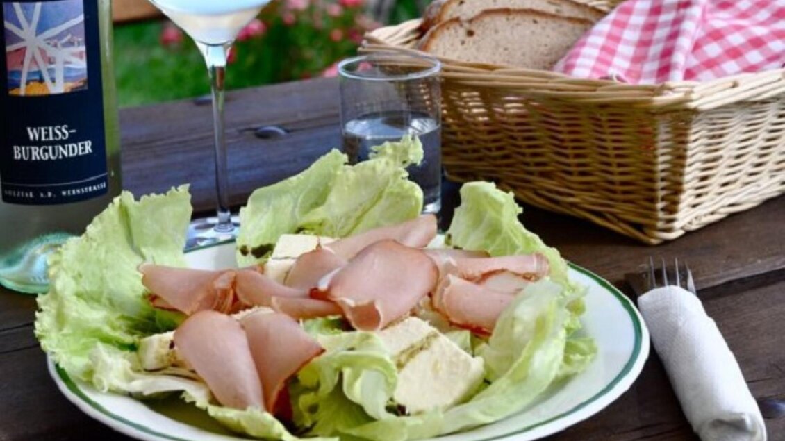 Ein Teller mit Salat, Aufschnitt und Käse, serviert auf einem Holztisch. Im Hintergrund stehen ein Glas Wein und ein Korb mit Brot. | © Trunk