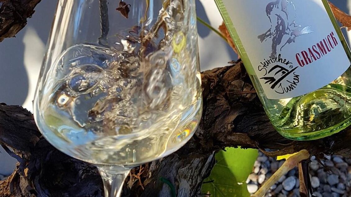 Ein Glas Weisswein mit sprudelndem Inhalt steht neben einer Flasche mit dem Etikett „Grasnüg“. Im Hintergrund sind Weinreben und Kieselsteine zu sehen. | © Grasmuck