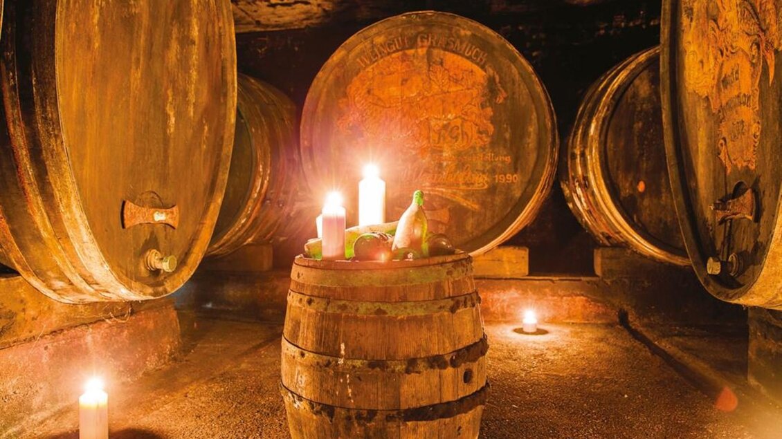 Ein stimmungsvolles Weinkeller-Interieur mit großen Holzfässern und einem Holzfass als Tisch. Kerzenlicht sorgt für eine gemütliche Atmosphäre. | © Grasmuck