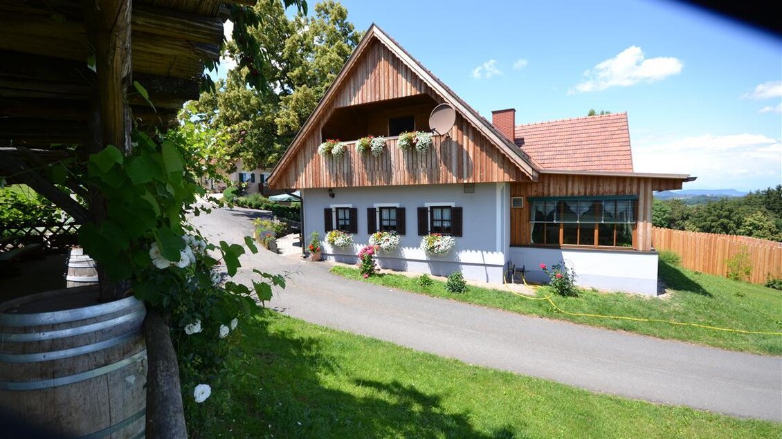 Ein hübsches Haus mit Holzfassade und Blumenbalkonen steht in einer grünen Umgebung. Im Vordergrund ist ein Weg, der zum Haus führt. | © Adam Schererkogl