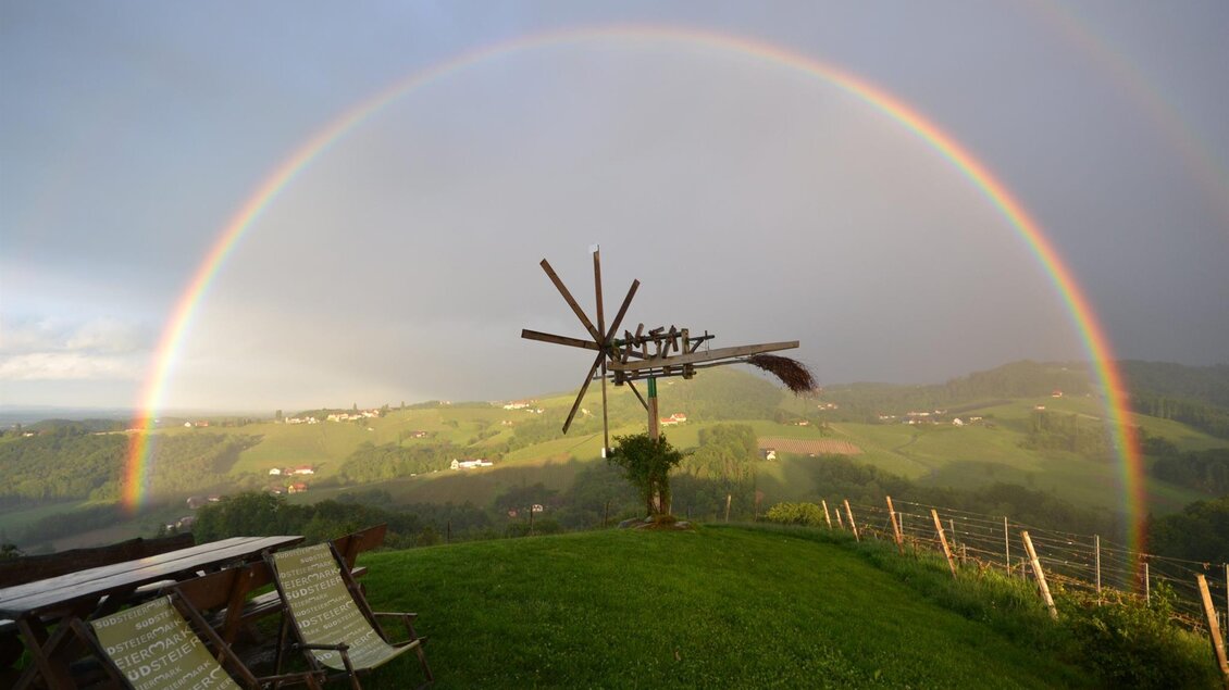 Ein schöner Regenbogen spannt sich über eine grüne landscape. Im Vordergrund sind Stühle und ein Windrad sichtbar. | © Adam