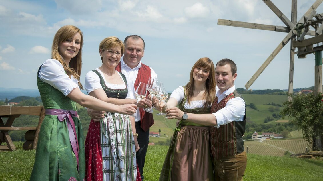 Eine Gruppe von fünf Personen in traditioneller Festkleidung hebt Gläser zum Anstoßen. Im Hintergrund sind sanfte Hügel und ein Windrad sichtbar. | © Adam