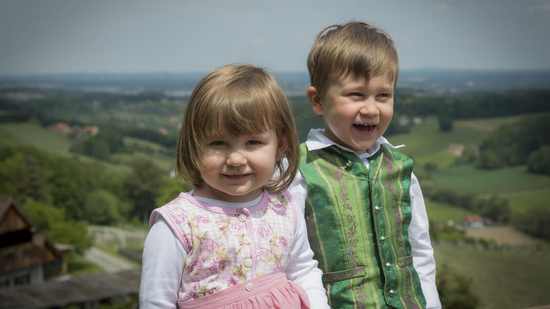 Zwei fröhliche Kinder stehen in einer wunderschönen Landschaft. Im Hintergrund sind grüne Hügel und ein blauer Himmel zu sehen. | © Adam